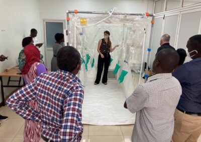 A group of healthcare professionals stand around a large clear plastic isolation enclosure while one person stands inside the enclosure demonstrating or explaining its features in a clinical room.