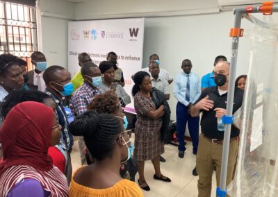 A group of participants stand closely together indoors while listening to a presenter who gestures toward a clear plastic isolation enclosure, with a banner and training materials visible in the background.