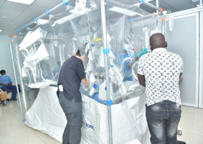 Patient lies in a hospital bed inside a large clear plastic isolation enclosure while multiple healthcare workers wearing protective masks and gloves provide care from outside and through access openings, with additional staff seated nearby in the background.