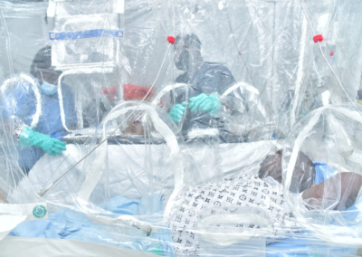 Patient lies in a hospital bed inside a clear plastic isolation enclosure while multiple healthcare workers wearing protective gowns, masks, and gloves provide care through built‑in access ports.
